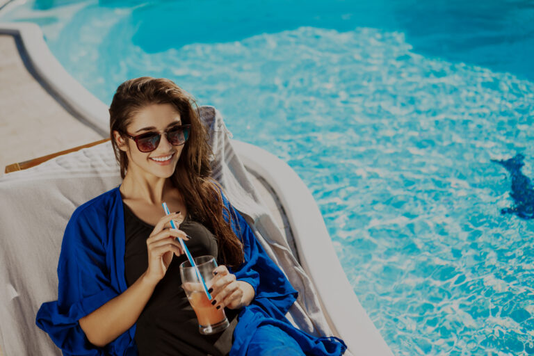 Beautiful brunette girl smiling, drinking cocktail, lying on chaise near swimming pool. Copy space.